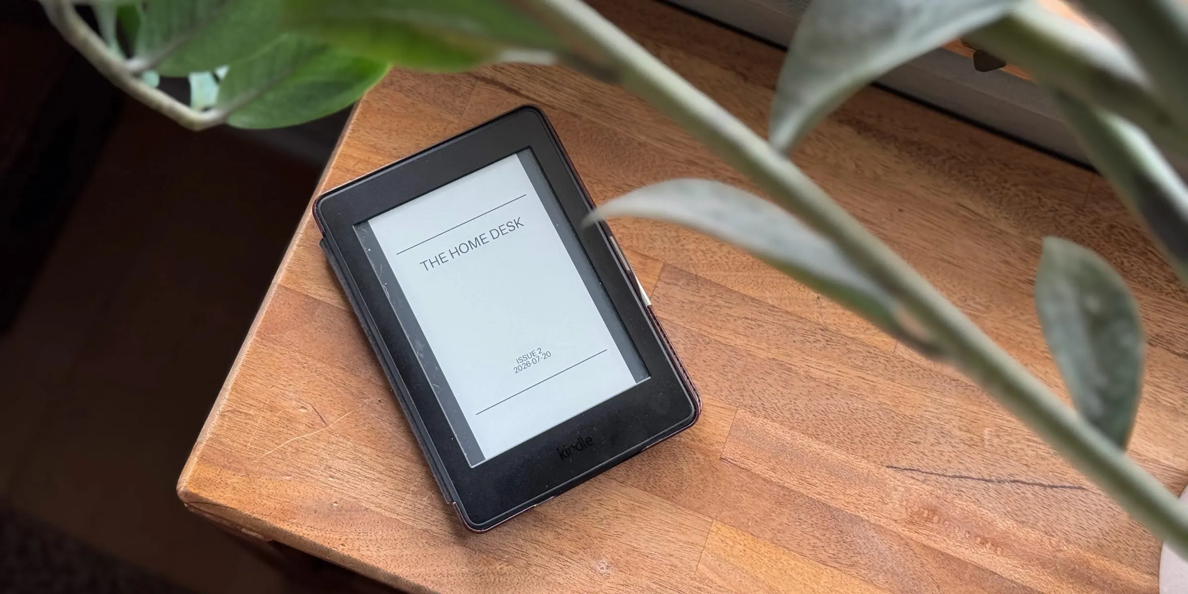 A Kindle on a wooden side table, framed by houseplant leaves, showing the plain cover of The Home Desk — Issue 2, dated 2026.07.20.